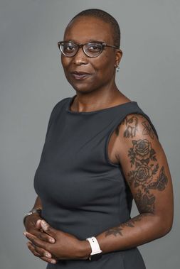 Lupe Davidson smiles softly for the camera while wearing glasses, a black, sleeveless dress that shows flower tattoos down her arm. 